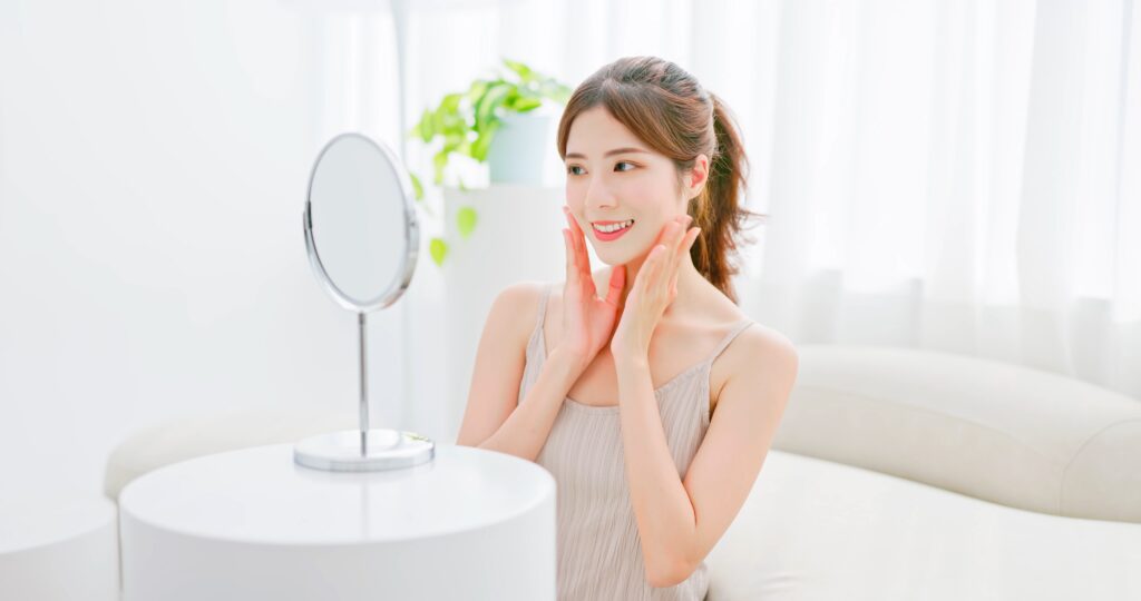 Eine junge asiatische Frau blickt zufrieden in einen Handspiegel und freut sich über das Ergebnis ihrer Beauty-Behandlung zuhause.